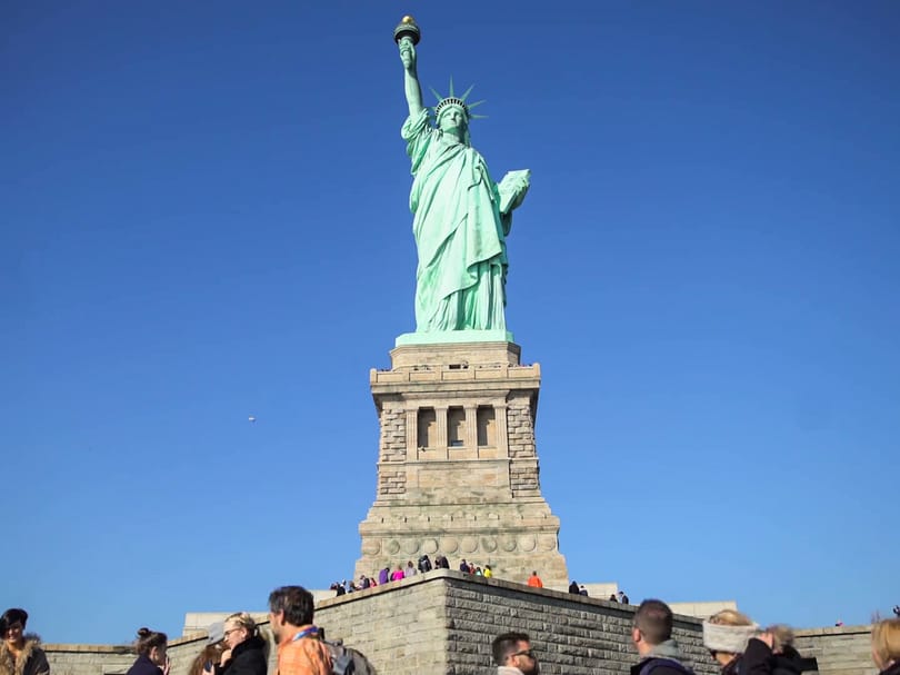 Full view of the Statue of Liberty and its pedestal on Liberty Island under a clear blue sky, with visitors gathered below.