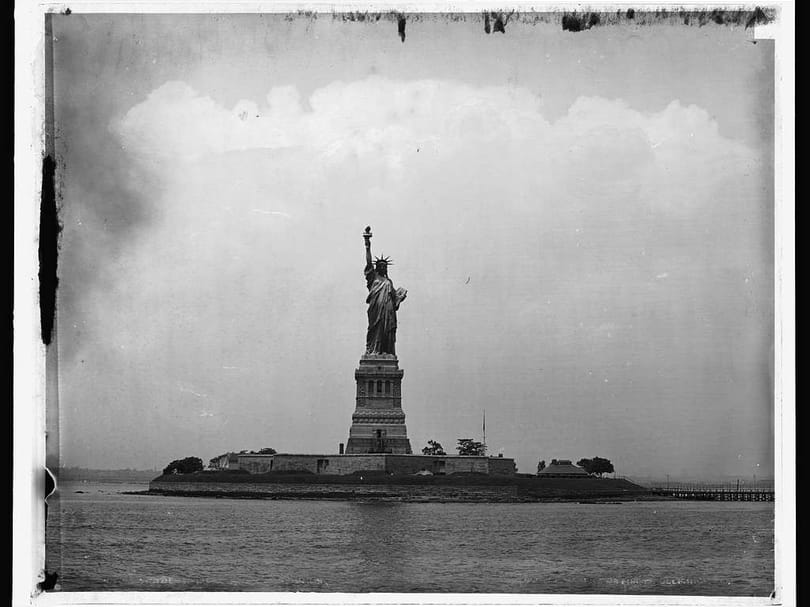 Historic black-and-white photograph of the Statue of Liberty on Liberty Island with the Manhattan skyline visible across New York Harbor.