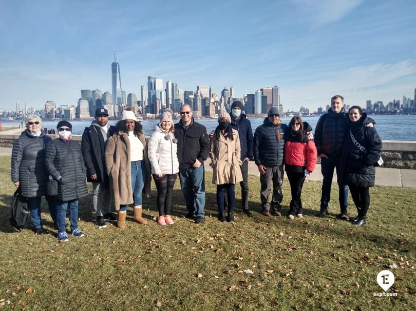 Group of visitors posing on Liberty Island with the Lower Manhattan skyline and One World Trade Center in the background.