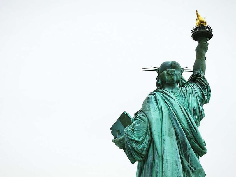 Rear view of the Statue of Liberty showing her raised torch, crown, tablet, and copper exterior against an overcast sky.