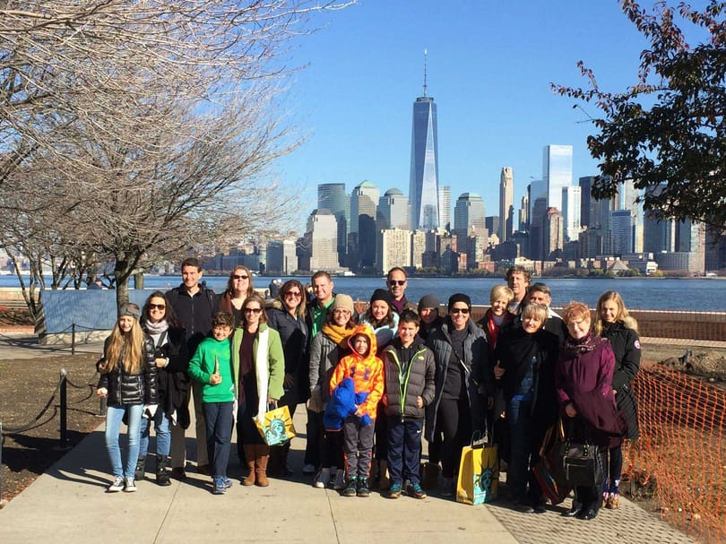 Group of visitors posing near the waterfront with the Lower Manhattan skyline and One World Trade Center behind them.